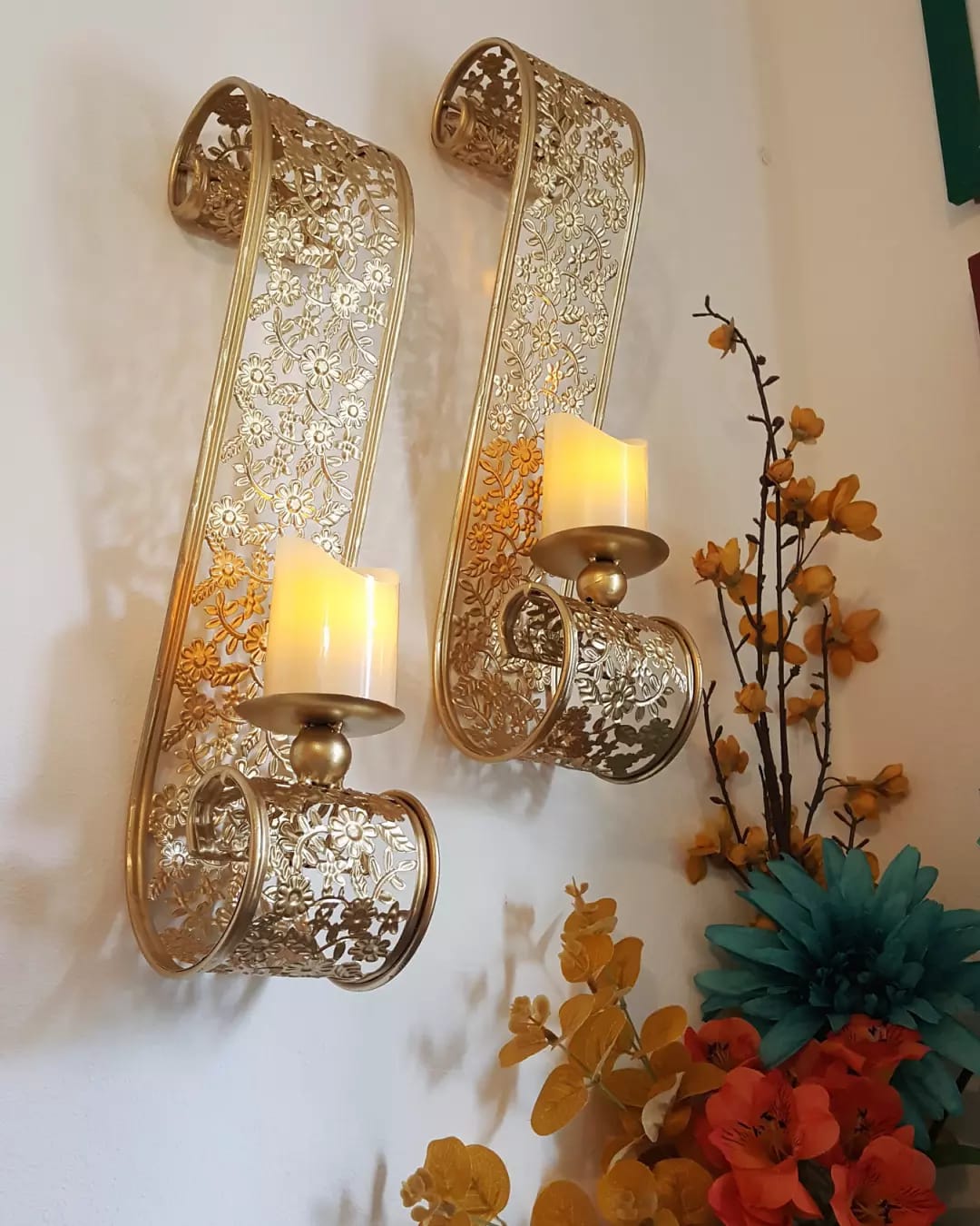 Pair of gold metal scroll wall candle sconces with lit candles beside flowers